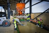 Drie mannen in gele veiligheidsoveralls. Een man bedient een oranje betonkubel. Twee andere mannen strijken het gestorte beton glad met planken.