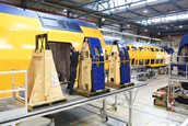 Geelblauwe dubbeldekkertrein van NS, met daarvoor een steiger met daarop drie houten stellages. Op twee van de stellages staan twee blauwe deuren met bij een van de stellages een man in donderblauwe werkkleding.